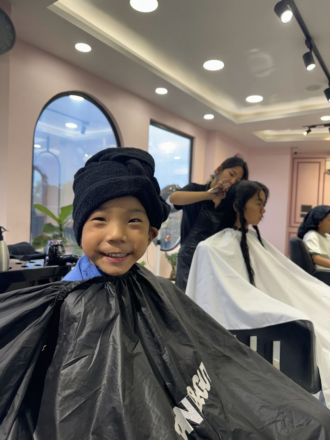 A smiling young boy in a beanie and cape waiting for his haircut.